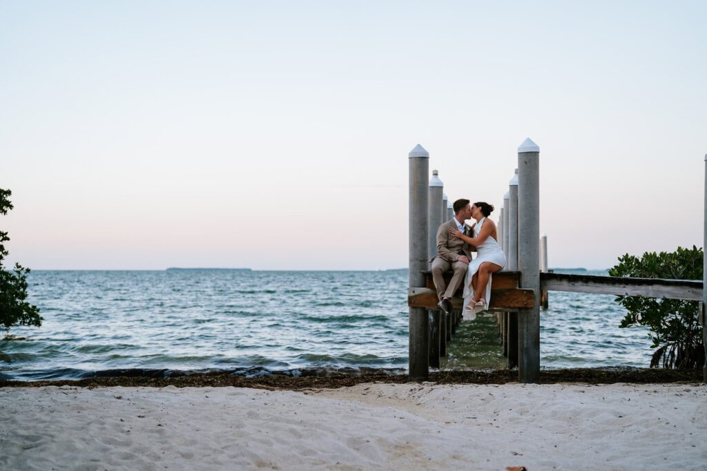 bride and groom sitting on a pier, kissing, during their golden hour wedding portrait session in Key Largo, Fl