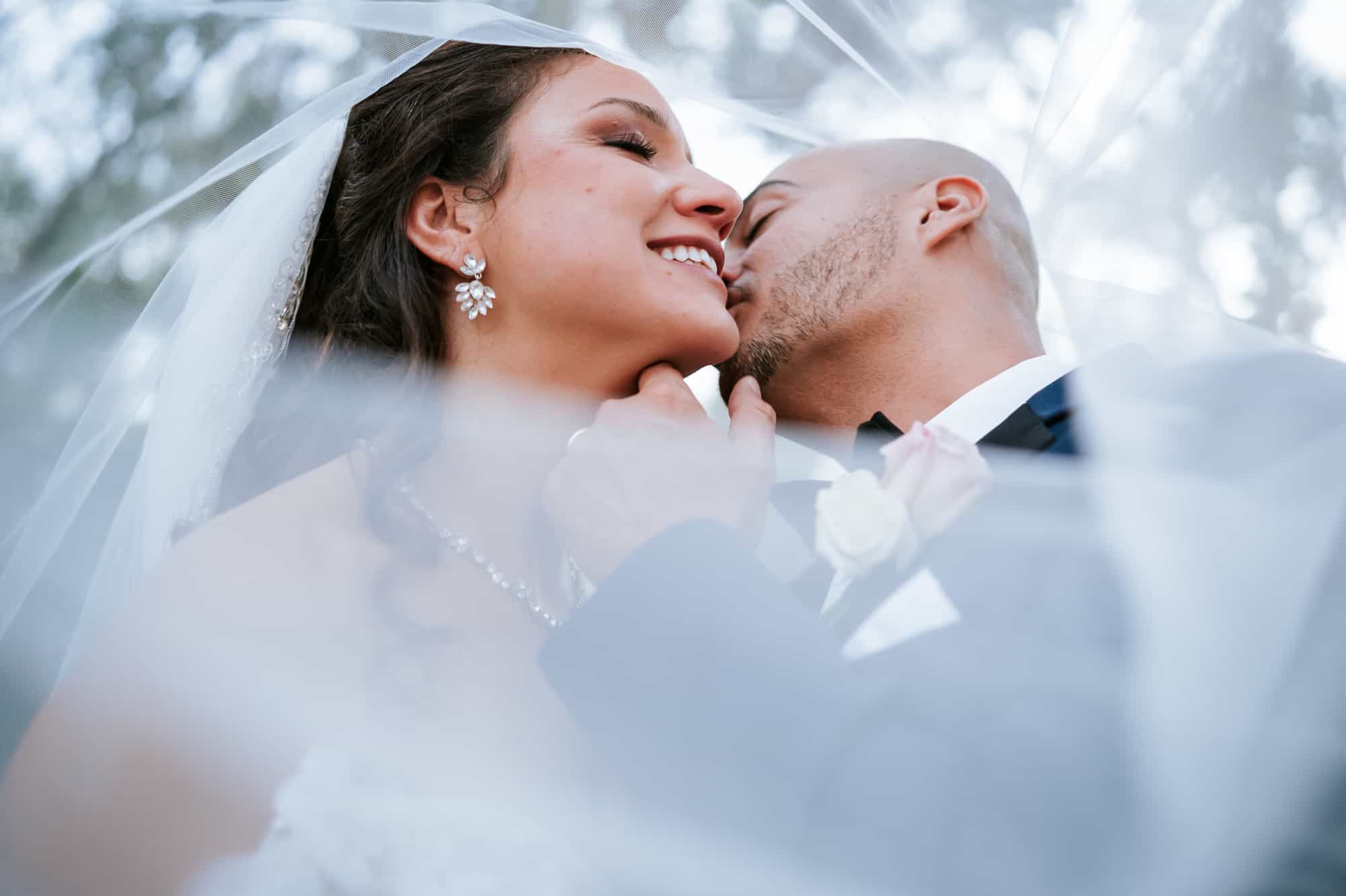 Wedding portrait of a veil shot with the groom kissing the bride's cheek on their wedding day at Bakers Ranch Wedding Venue