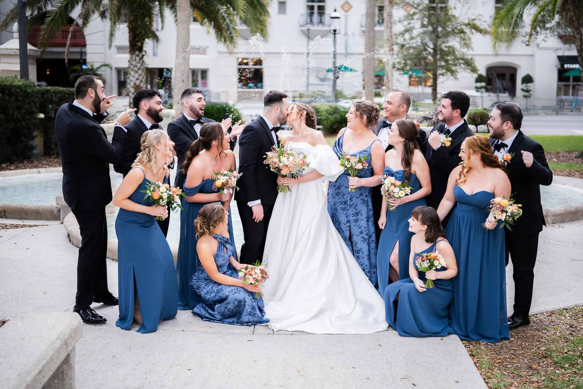 The bride and groom share a kiss surrounded by a large wedding party in the Casa Monica courtyard, with bridesmaids in slate blue and groomsmen in black tuxedos captured by Atlanta Wedding Photographer Peach State Weddings