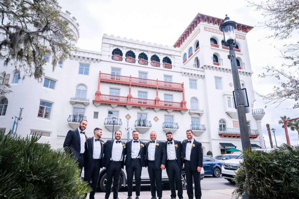 Seven groomsmen in black tuxedos with bow ties stand together in a courtyard at the Casa Monica Resort.