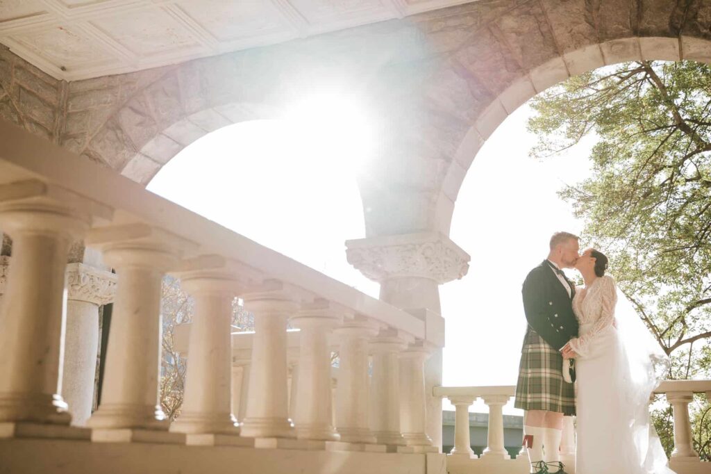 Intimate moment between a bride and groom in a kilt standing beneath the Romanesque arches of a historic Georgia mansion.
