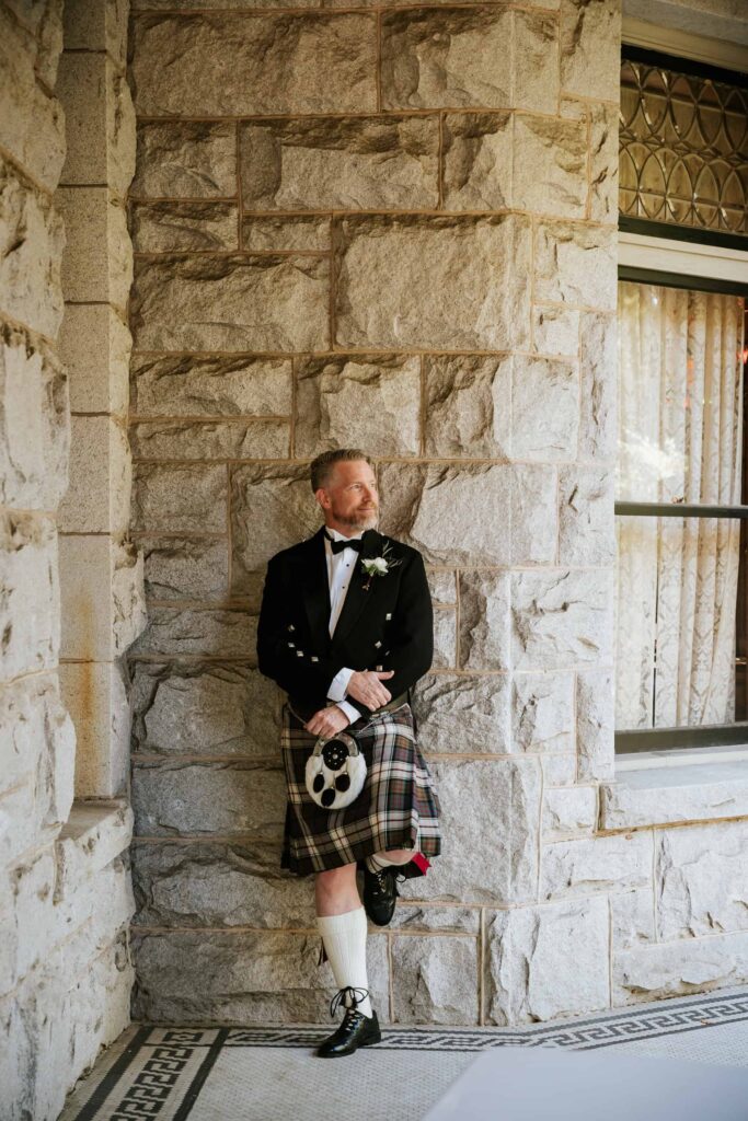Groom in a full Prince Charlie jacket and kilt leaning against the historic stone walls of an Atlanta castle venue.