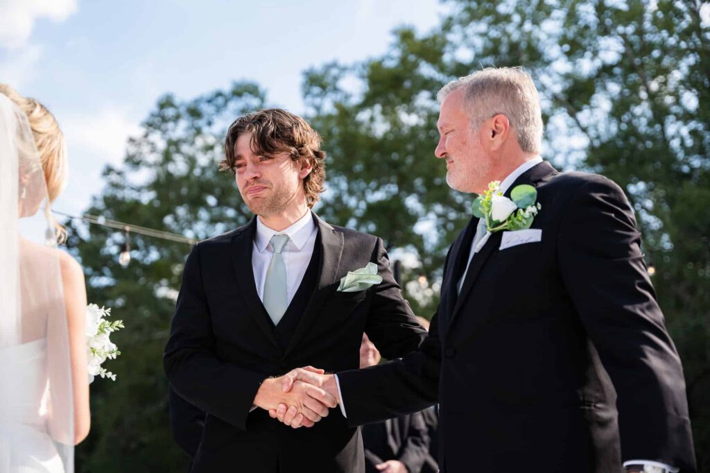 Portfolio 3 candid moment of the father of the bride shaking the grooms hand during the wedding ceremony. groom is crying while he looks at his beautiful bride at The Venue at Daisy Hill in Fayetteville GA