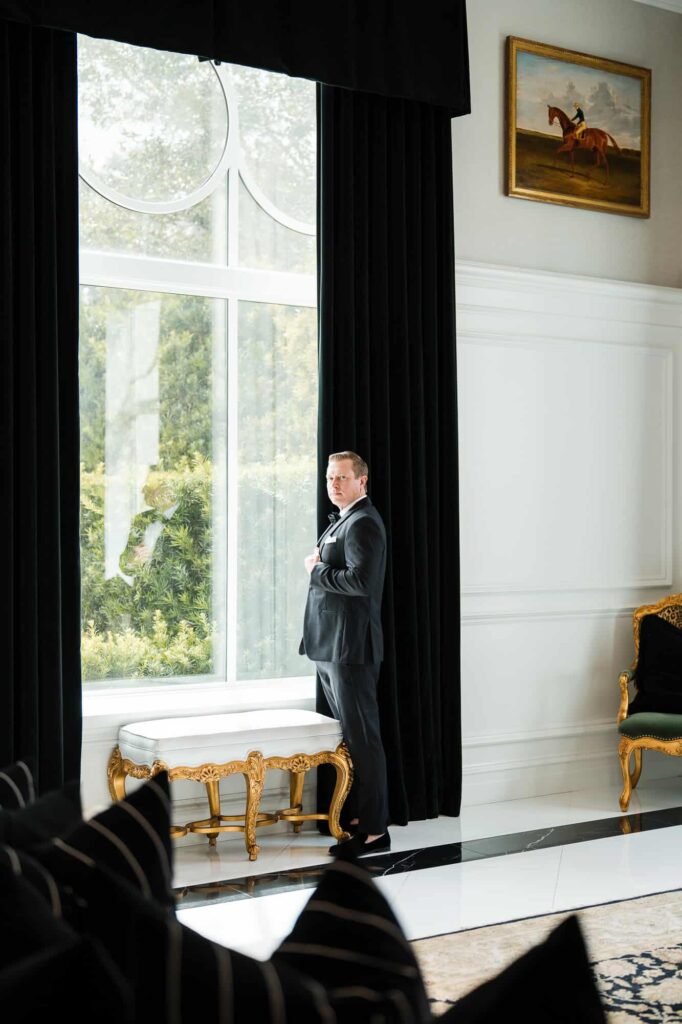 A high-end wedding portrait of a groom in a tuxedo, framed by large windows at the World Equestrian Center.