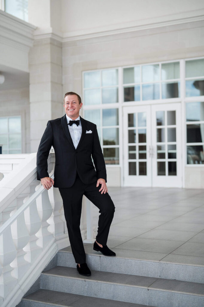 A groom in a black tuxedo leans against a white balustrade at the World Equestrian Center, captured by an Atlanta Georgia wedding photographer.